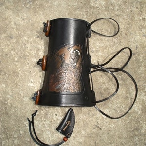 K&ouml;nnte beinhalten: Schwarzer Lederarmschutz mit einem braunen B&auml;renmotiv und einer kleinen Ledertasche mit einem Symbol.