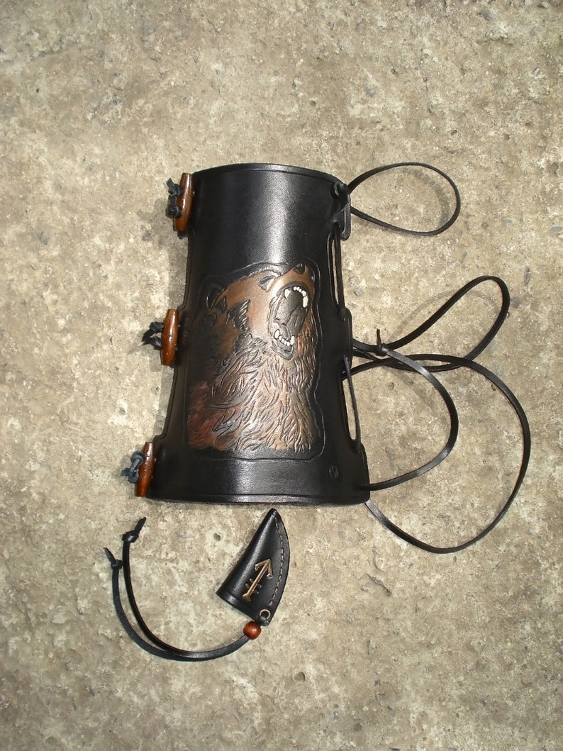 K&ouml;nnte beinhalten: Schwarzer Lederarmschutz mit einem braunen B&auml;renmotiv und einer kleinen Ledertasche mit einem Symbol.
