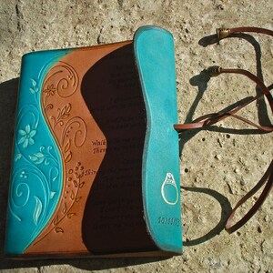 Op de afbeelding: Een bruin lederen dagboek met een turquoise blauwe flap en een bloemmotief. Het dagboek heeft een leren band met een metalen sluiting en de woorden "Walk They Shine" zijn op de omslag gegraveerd.
