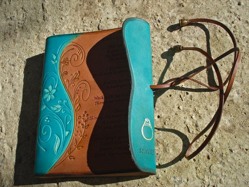 Op de afbeelding: Een bruin lederen dagboek met een turquoise blauwe flap en een bloemmotief. Het dagboek heeft een leren band met een metalen sluiting en de woorden "Walk They Shine" zijn op de omslag gegraveerd.