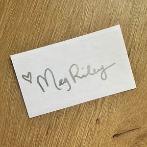 May include: A white card with a silver heart and the handwritten signature "Meg Riley".