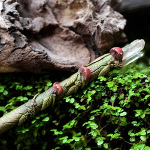 Może przedstawiać: Ręcznie wykonana różdżka z przezroczystą kryształową końcówką, ozdobiona czerwonymi akcentami grzybów i zielonymi detalami liści. Różdżka ma około 30 cm długości, spoczywa na zielonych liściach.