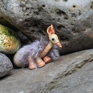 May include: A small, sculpted creature figurine with a smooth, light beige head, pointed ears with reddish-pink inner colouring, and a long neck. The body is covered in long, soft, greyish-purple faux fur, and it has rounded, textured feet.
