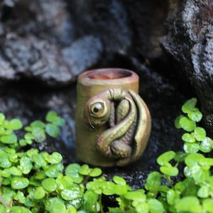 May include: A cylindrical, olive-green ceramic bead featuring a sculpted, coiled creature with a prominent eye and textured skin wrapped around the exterior surface. The bead has a hollow center and a smooth, matte finish on its outer body.