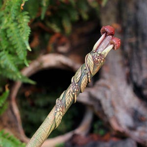 May include: A decorative wand featuring a textured green shaft with sculpted leaves and small berry clusters winding around it. The top of the wand is adorned with two small, rounded, reddish-brown mushroom caps emerging from a leafy base.