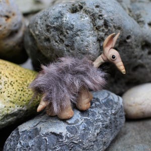 May include: A small, handcrafted creature figurine with a smooth, light tan head and snout, large pointed ears, and a dark eye. The body is covered in long, shaggy, greyish-purple faux fur, and the creature has visible, rounded, light tan feet.
