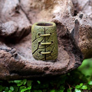 May include: A cylindrical bead with a textured olive green surface resembling cracked leaf veins. The bead features a vertical seam down the center with three horizontal light-colored stitches crossing the seam, creating a laced appearance.