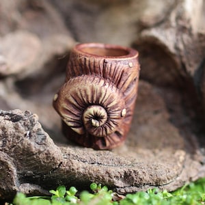 May include: A brown, handcrafted ceramic miniature snail shell planter. The shell features detailed spiral and ribbed textures, with a small opening at the top for planting. The planter is set on a natural stone surface.