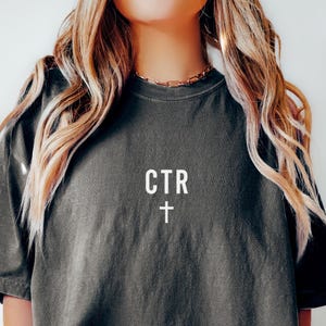 Op de afbeelding: Donkergrijs T-shirt met de tekst "CTR" en een wit kruis. Het shirt heeft een relaxte pasvorm en korte mouwen. Het model draagt een grijze short.