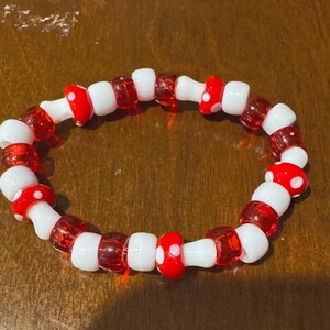 Puede incluir: Un brazalete de cuentas rojas y blancas con cuentas con forma de hongo. El brazalete está hecho de pequeñas cuentas redondas que se ensartan para formar un círculo.