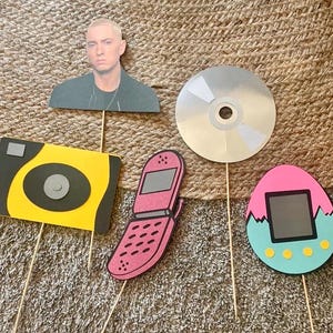 Puede incluir: Una colección de recortes de papel sobre palitos de madera, que incluye un retrato, un CD, una cámara, un teléfono plegable y un huevo estilo Tamagotchi. Los artículos son de varios colores, incluyendo amarillo, rosa y azul.