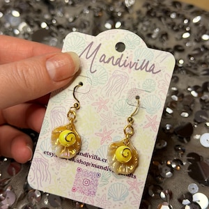 May include: A pair of gold-tone hook earrings featuring a small, iridescent seashell charm with a bright yellow bead at the centre. The earrings are attached to a branded display card with the text Mandivilla and a QR code, decorated with a light-coloured pattern of seashells and starfish.