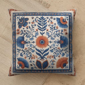 May include: A square throw pillow featuring a symmetrical floral pattern in shades of blue and burnt orange on an off-white background. The design includes a central sun-like flower surrounded by stylized leaves, vines, and blossoms.