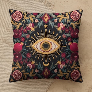 May include: A square throw pillow with a black background featuring a central embroidered golden eye with radiating sunburst rays and a crescent moon below. The design is surrounded by a botanical pattern of red pomegranates and green leaves.