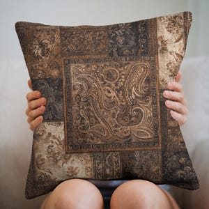 May include: Square throw pillow with a patchwork design in shades of brown, tan, and black. The center panel features a detailed paisley pattern, while the surrounding squares showcase floral and geometric motifs. The pillow is held by a person.