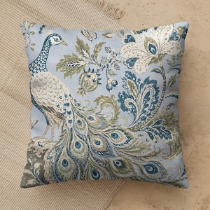 May include: A square throw pillow featuring a light blue background with a detailed peacock illustration. The design includes intricate floral and botanical patterns in shades of dark blue, olive green, and cream, covering the entire surface.