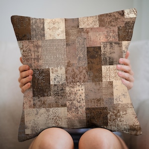 May include: Square decorative pillow with a patchwork design in shades of brown, beige, and taupe. The pillow features a variety of textures and patterns, including floral and geometric motifs. The pillow is held by a person.