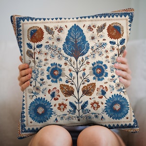 May include: Square decorative pillow with a floral pattern in shades of blue, brown, and cream. The design features stylized flowers and leaves, with a detailed border. The pillow is held by a person.