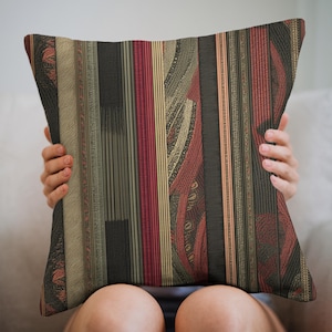May include: Decorative square pillow with a vertical striped pattern in shades of red, black, gold, and olive green. The pillow has a textured appearance, with a mix of solid and patterned stripes. The pillow is being held by a person.