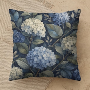 May include: A square throw pillow featuring a botanical print of hydrangea flowers in shades of white, light blue, and deep navy. The pattern includes detailed green and blue leaves set against a dark navy background, creating a floral design.