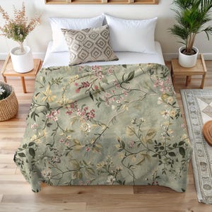 Puede incluir: Una manta con estampado floral en tonos verdes, rosas y crema, sobre una cama. La manta presenta un delicado diseño botánico de flores y hojas. La cama está hecha con almohadas blancas y un cojín estampado.