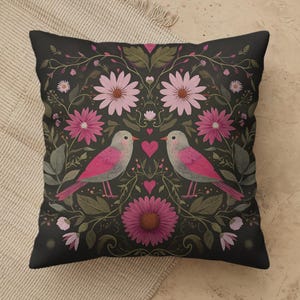 May include: A square throw pillow featuring a dark charcoal background with a symmetrical floral pattern. Two birds with pink and gray plumage face each other, surrounded by pink daisies, green leaves, and small pink hearts in a decorative arrangement.