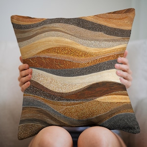 May include: Decorative throw pillow with a layered, abstract design in shades of brown, beige, and tan. The pillow features a textured pattern of small dots and wavy lines, creating a visual depth. The pillow is held by a person.