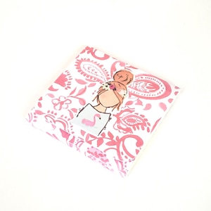 May include: A square, folded fabric with a pink paisley and floral pattern. A sticker of a woman with a bun and a flamingo on a sign is on top. The background is white.