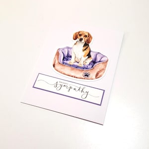 May include: A sympathy card featuring a watercolor illustration of a beagle dog sitting in a purple and brown dog bed. The card includes the words "With deepest sympathy" in elegant script within a purple border.