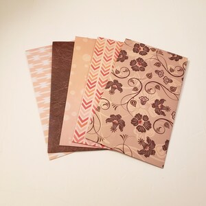May include: Set of five decorative paper sheets with different patterns. The patterns include a geometric design, a brown leather texture, a polka dot pattern, a chevron pattern, and a floral design. The sheets are all in shades of brown, pink, and white.