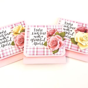 May include: Three pink and white plaid cards with the text "Start each day with a grateful heart." Each card is decorated with a pink, yellow, or red rose and green leaves. The cards are on a white background.