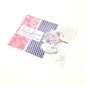May include: A greeting card with a patchwork design. The card features pink floral patterns, blue and white gingham, and a floral illustration with a teacup and bird. The text "delight in the little things" is printed on the card.