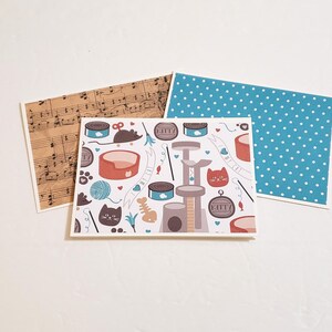 May include: Three greeting cards with white borders. One card features a musical score, another has a blue polka dot pattern, and the central card displays a cat-themed design with cat toys, food, and a cat tree.