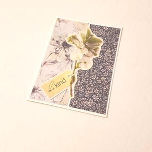 May include: A greeting card with a floral design. The card features a light floral pattern on the left and a dark floral pattern on the right. A sprig of white flowers and a tag that says "be kind" are attached to the card.