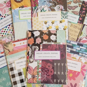 May include: A collection of decorative paper packs with various patterns and colors. Designs include floral, butterfly, bee, musical notes, and geometric patterns. Each pack has a label that reads "DREAM. CREATE. INSPIRE." and a website address.