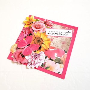 May include: A handmade greeting card with a vibrant floral arrangement. The card features a pink border and a collage of flowers, including roses, sunflowers, and a butterfly. The words "special moments" are printed on a white rectangle.