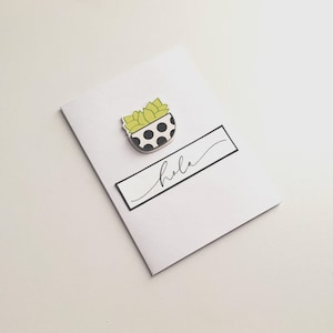 May include: A white card with a black rectangle containing the word "hola" in cursive. A small, decorative succulent in a black and white polka-dot pot is affixed to the card. The succulent has green leaves.