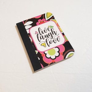 May include: A floral patterned notebook with the words "live laugh love" in a white and pink frame. The notebook has a black spine and a colorful design with pink, green, and brown flowers.