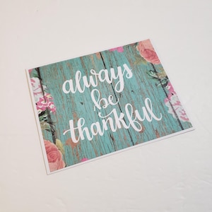 May include: A square card with the words "always be thankful" in white script on a turquoise wood-grain background. Pink and white floral accents frame the edges of the card, creating a decorative and inspirational design.