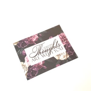 May include: A sympathy card with a floral design in shades of purple, white, and black. The card features the text "Thoughts are with you" in elegant script.