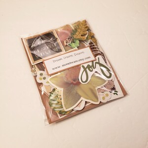 May include: A clear plastic bag filled with various paper craft supplies, including stickers, die cuts, and embellishments. The items are in shades of brown, green, and gold. The bag also includes a small card with the text "Dream. Create. Inspire. www.monetware.etsy.com" and a large die cut with the word "joy" in green lettering.