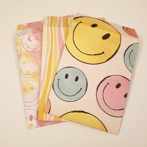 May include: Three decorative paper bags with colorful designs. One bag features a pattern of yellow smiley faces on a pink background. Another has vertical stripes in shades of yellow and orange. The third bag has a pattern of smiley faces, hearts, and stars.