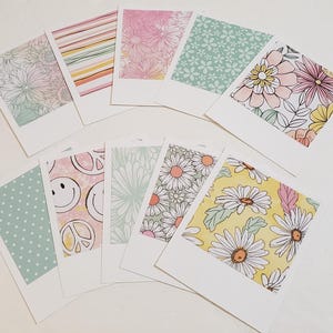 May include: A collection of nine polaroid-style cards with various floral and patterned designs. The cards feature pastel colors, including pink, yellow, and green, with daisy, stripe, and polka dot patterns. The cards are arranged on a white surface.