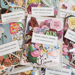 May include: A collection of colorful paper die cuts and stickers, including butterflies, flowers, and inspirational quotes. Some packages have the text "DREAM. CREATE. INSPIRE." and a website address. The stickers feature a flamingo, a bunny, and the words "Be a Queen."