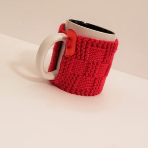 May include: A red knitted mug cozy with a white ceramic mug. The cozy has a woven pattern and is designed to keep drinks warm.
