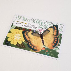 May include: A greeting card with a large butterfly and the words "happy ANNIVERSARY". The butterfly has orange and black wings. The card has a floral design with yellow flowers and green leaves.