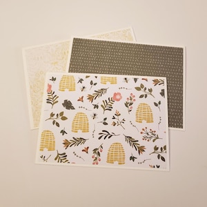May include: A collection of decorative greeting cards. One card features a white background with a pattern of gold beehives, leaves, and flowers. Another card has a dark green dotted pattern, and a third card has a gold floral design.