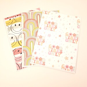 May include: Three decorative paper sheets with retro designs. One features smiley faces, another has rainbow arches, and the third displays the text "GOOD VIBES ONLY" with floral and star accents. The color palette includes pink, yellow, and peach.
