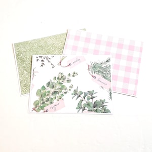 May include: Three decorative square papers. The central paper features watercolor illustrations of herbs like oregano and rosemary, with pink tags. A green paisley patterned paper and a pink and white gingham paper are also visible. The papers are arranged on a white surface.