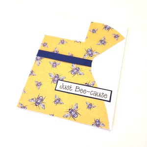 May include: A handmade greeting card with a yellow background covered in bee illustrations. A navy blue ribbon is across the card. The text "Just Bee-cause" is in a black rectangle.